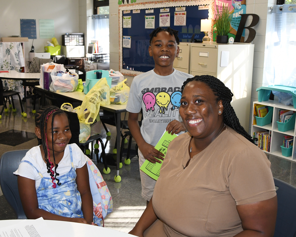 A classroom with three people. A woman in a brown shirt and two children, one with braids and one smiling.