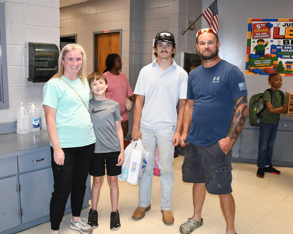 Three adults and two children in a room, two holding bags. One adult has a tattoo. A flag hangs on the wall.