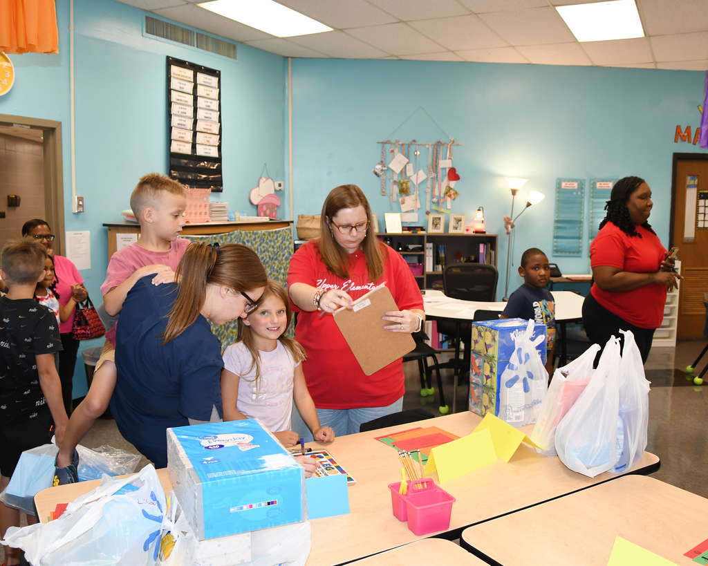 Group of children and adults in a classroom, examining boxes and items on a table.
