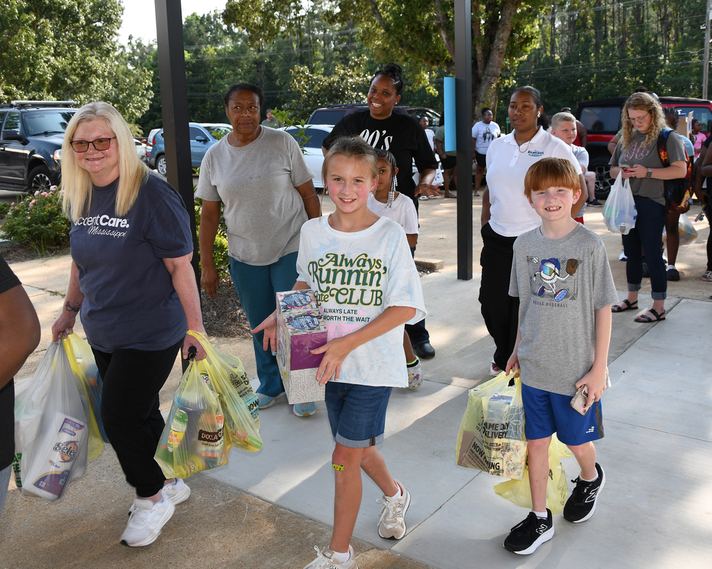 Group of adults and children holding bags, walking down a sidewalk in a parking lot.