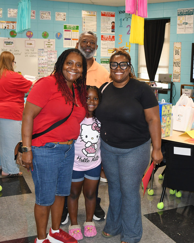 Three women and a girl in a classroom. One woman wears red, another black. The girl wears pink shoes.