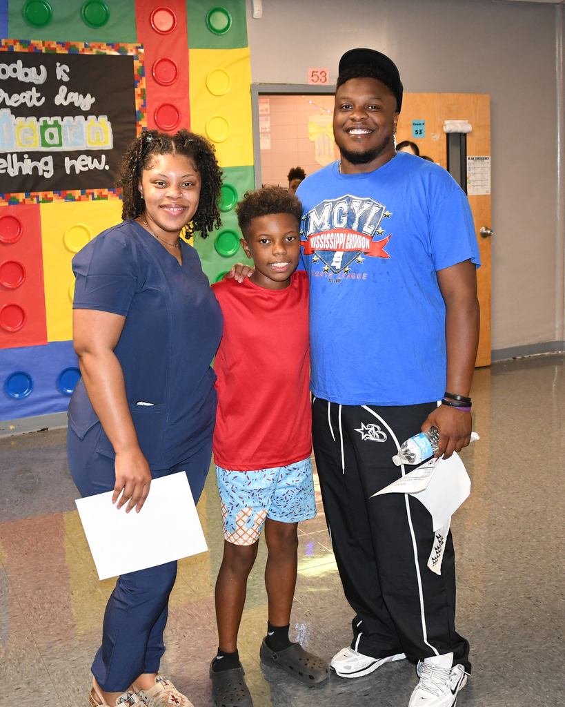 Three people stand in a corridor, smiling. A woman and man hold papers, and a boy stands between them. A colorful Lego wall is behind them.