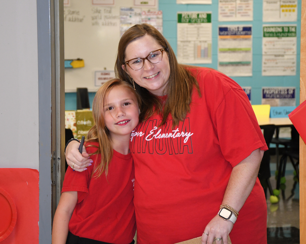 A woman and girl stand side by side, wearing red shirts with white letters. They are inside a room with blue walls, chairs, and a whiteboard.