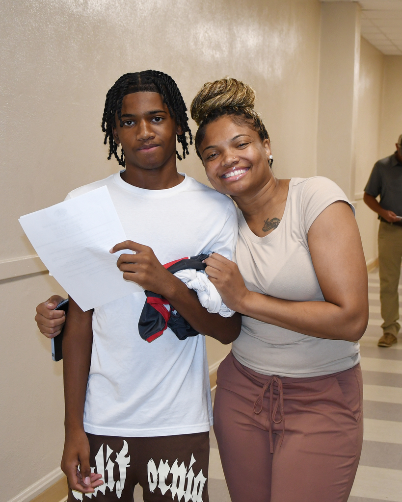 A boy and a woman stand close, smiling. The boy holds a paper, and the woman has a tattoo on her shoulder.