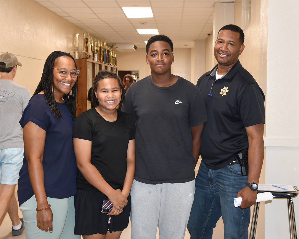 Four people in a hallway. Two men wear black shirts, one in jeans. Two women wear black tops, one with glasses. A trophy shelf is in the background.