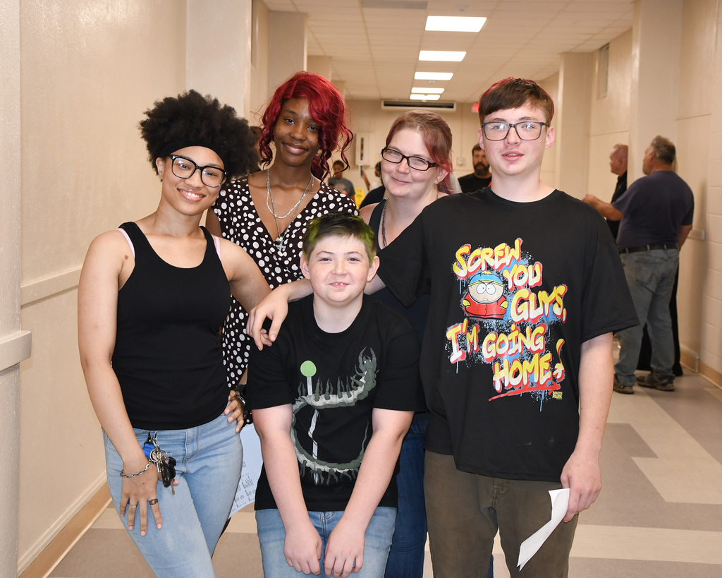 Five people stand in a hallway, smiling. A woman with curly hair and glasses is on the left.