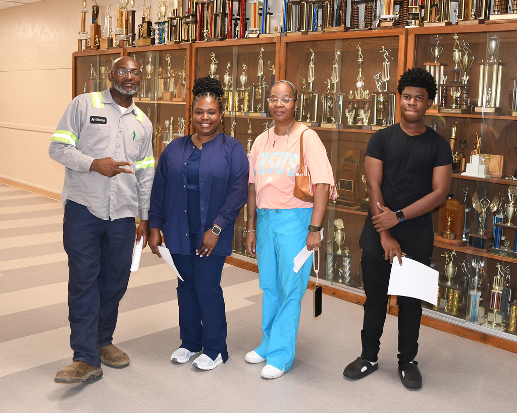 Four people stand in front of a trophy case, each holding papers. Two women wear pants, one in pink and one in blue. A man in gray stands to the left.