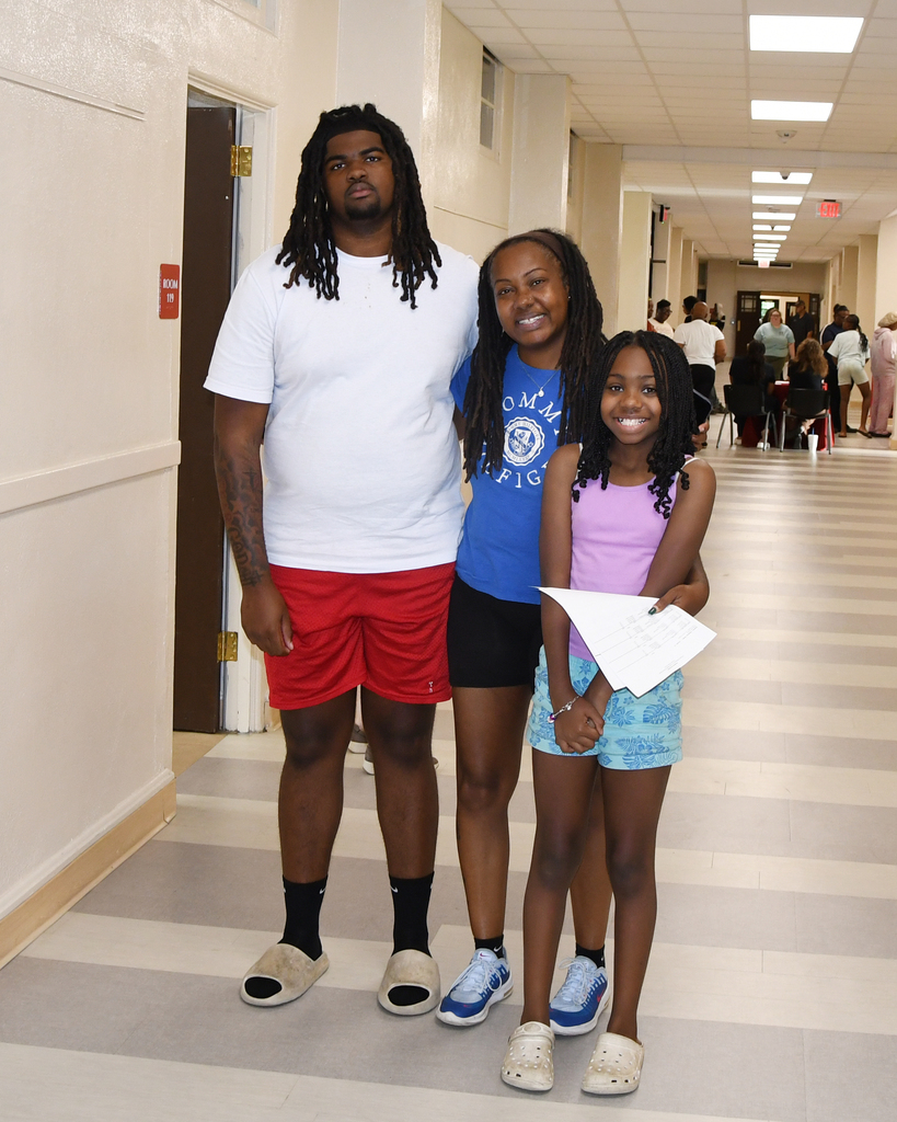 Three people, including a man, a woman, and a child, stand in a hallway with white walls and gray floors.