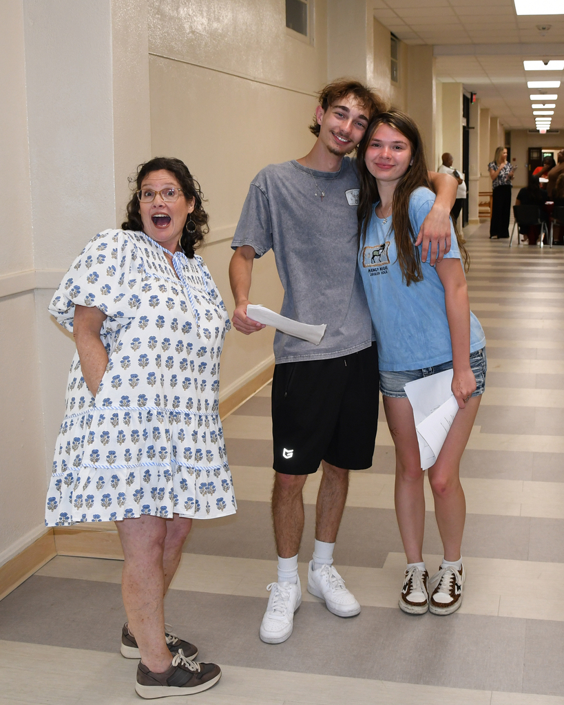 Three individuals stand in a hallway, smiling. The woman wears glasses, a floral dress, and sneakers. A man stands between two women, holding papers.