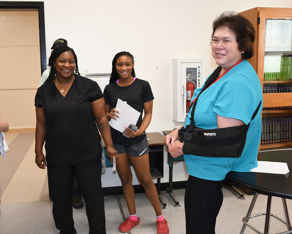 Three women in a room, one with a black sling. One woman holds a folder and speaks with the woman in blue.