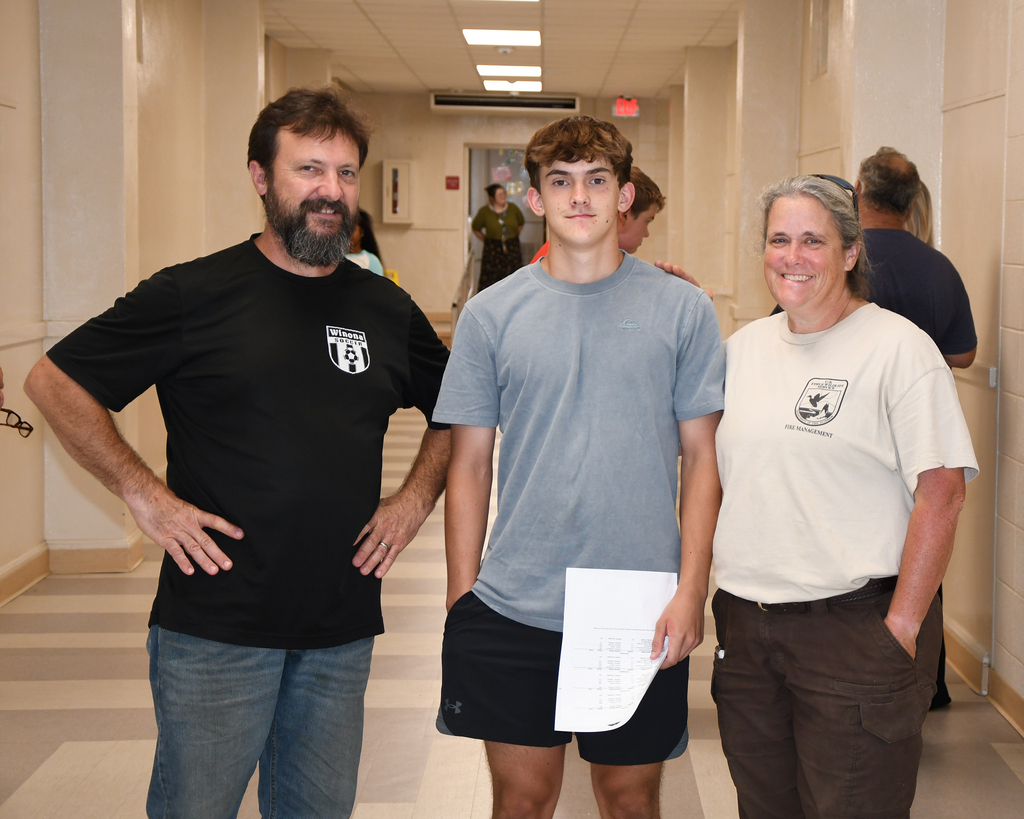 A man and woman stand with a boy in a school hallway. The boy holds a paper, while others walk behind them.