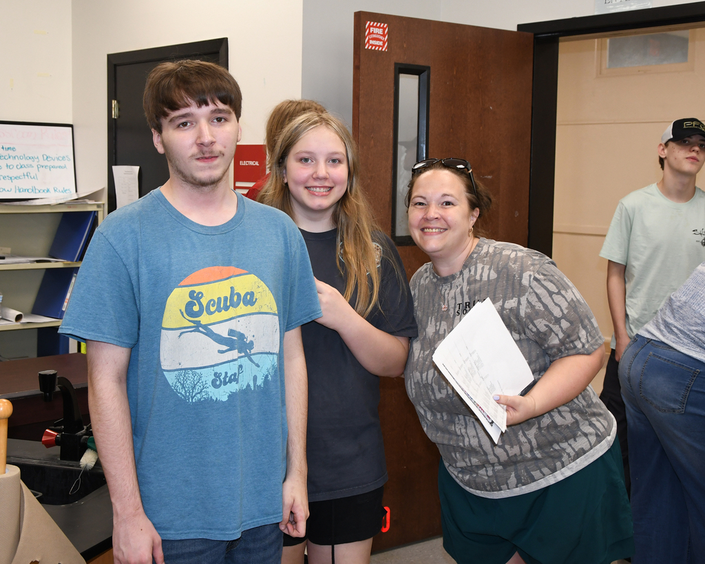 Four individuals stand in a room, two smiling. A woman holds papers. Behind them, a wooden door.