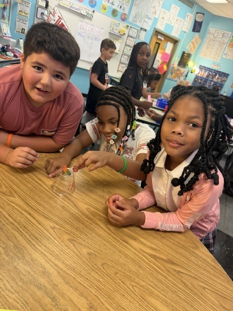 Three children sit at a table, two girls with braids and a boy with a smile, inside a classroom.