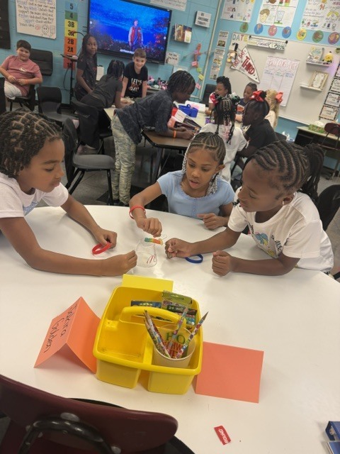 Children in a classroom sitting at a table. They are using scissors and other school supplies.