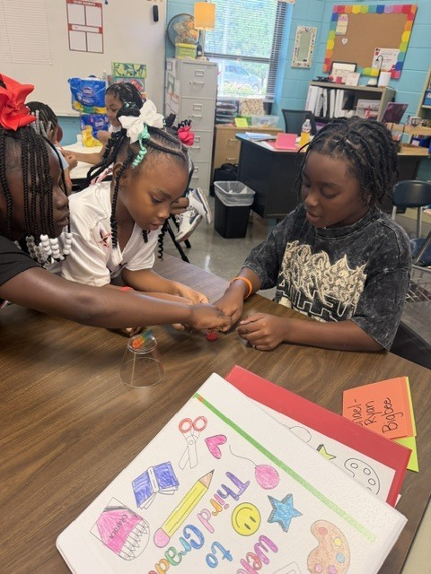 Three young girls sit at a table working on a craft project. One holds scissors.