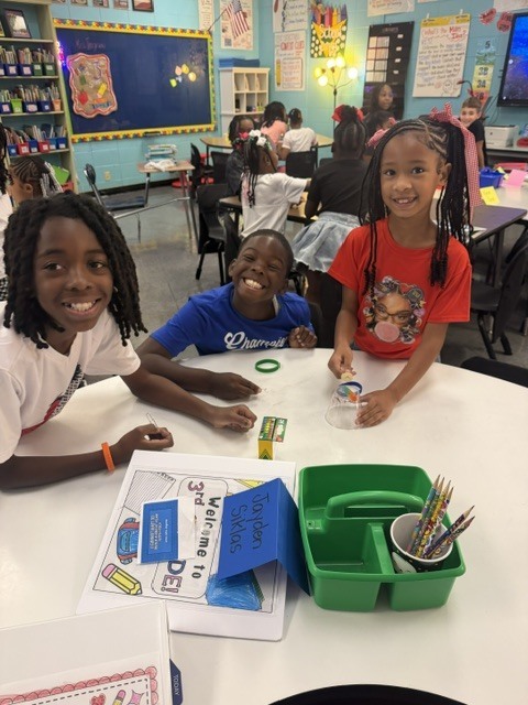 Three children sit at a table in a classroom, with one girl smiling and another holding a pencil.
