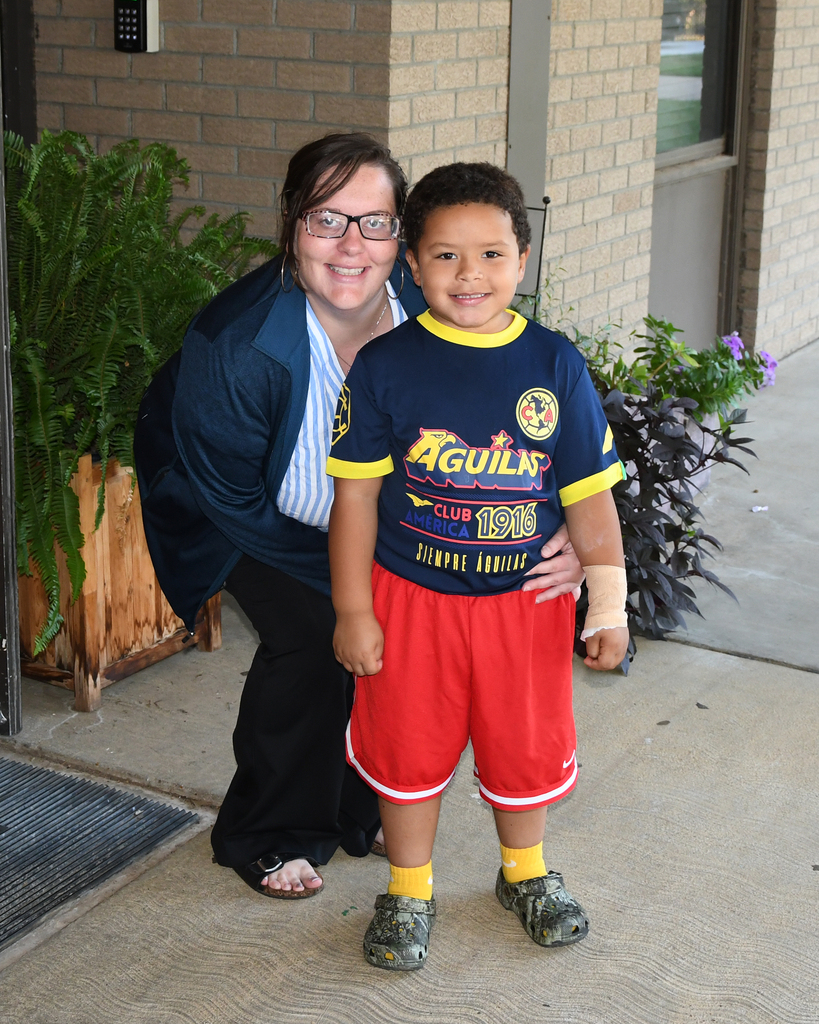 A woman and a child stand together on a concrete sidewalk in front of a brick building.