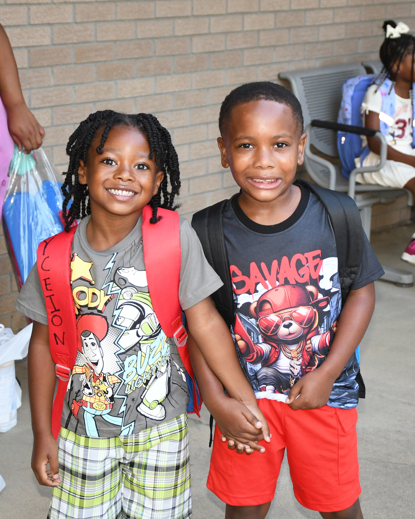 Two children smile for a photo, one wearing a red backpack, the other a black backpack with cartoon graphics.