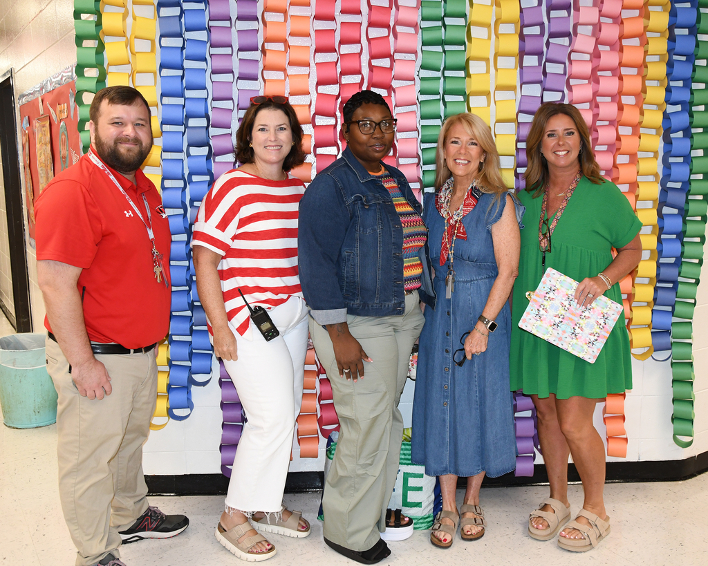 Five people stand in front of a multi-colored chain backdrop. One holds a paper bag.