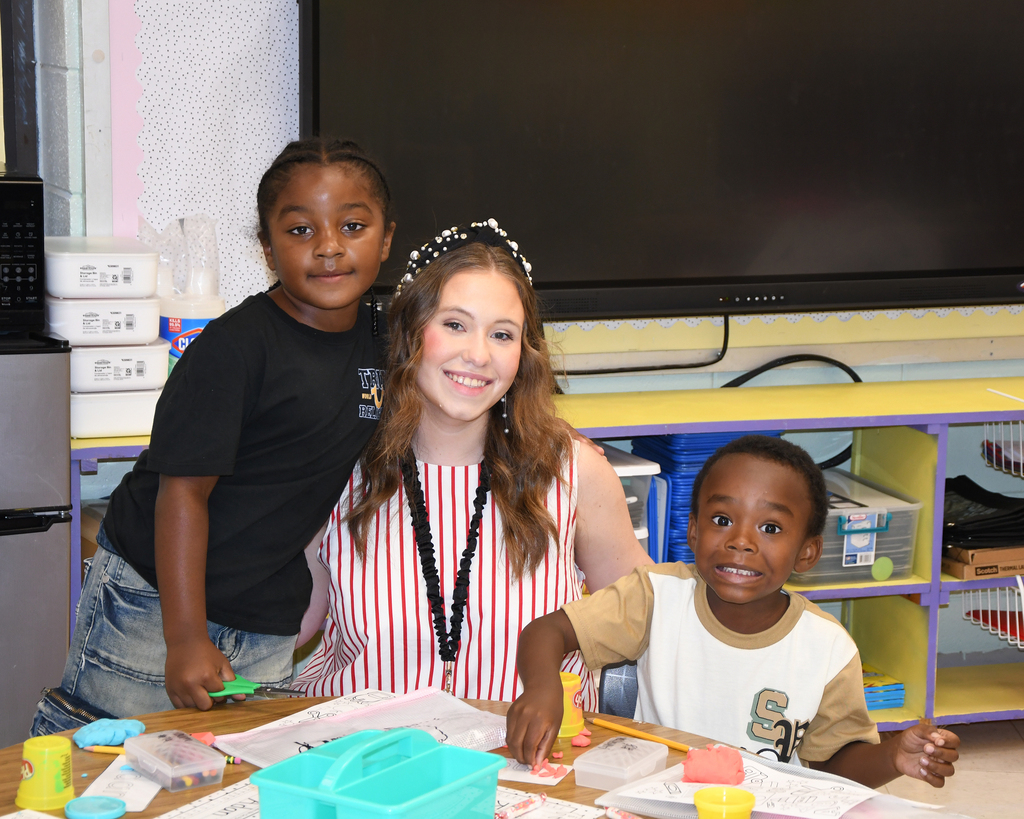 Three children in a classroom. A girl and boy sit at a table with craft materials. A girl stands behind them, smiling. A shelf with storage boxes and a monitor is behind them.