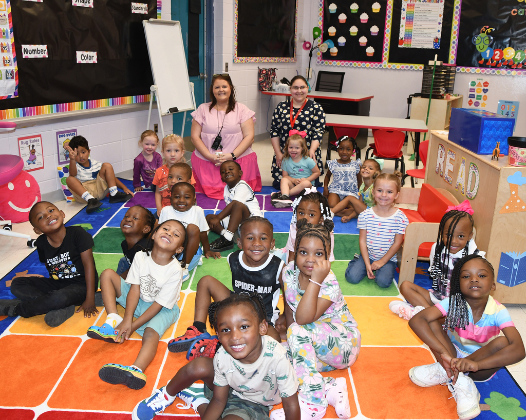 Classroom with children sitting on a colorful rug. Two adults stand near the back. Bright colors and cheerful faces.