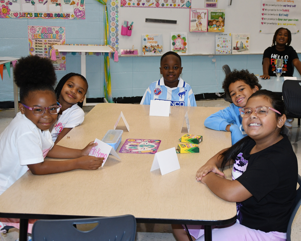 Five kids, all wearing glasses, seated at a table, smiling, with colorful materials in a classroom.