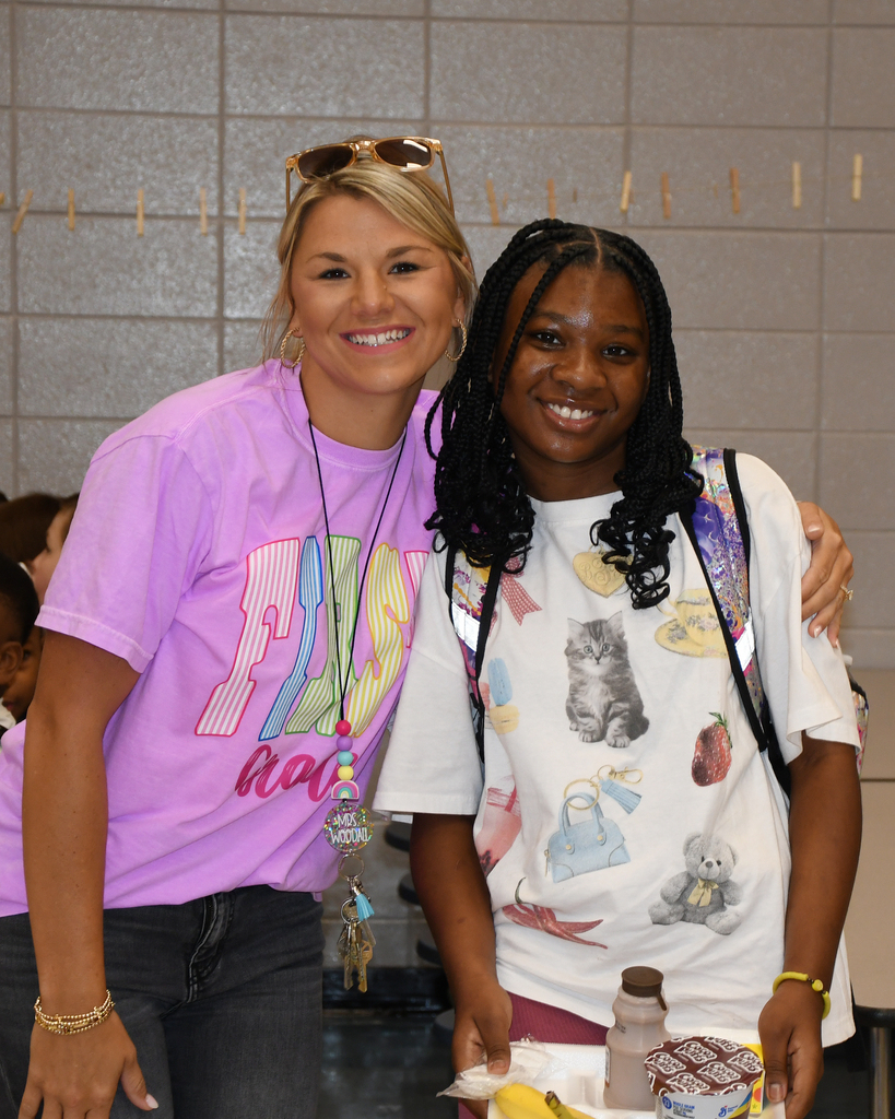 Two individuals stand next to each other, smiling, in a room with tiled walls. One wears a white shirt with various printed items.