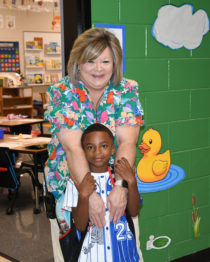 Woman and boy standing close together in front of a classroom wall with a duck painting.