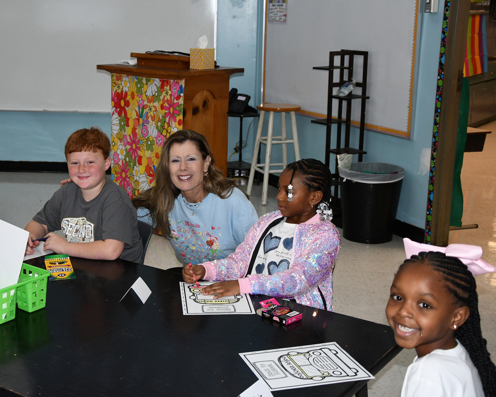 Three children and an adult woman sit at a table in a classroom, engaging with various materials.