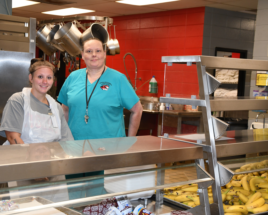Two women, one with a white apron and the other in a teal shirt, stand in a kitchen.