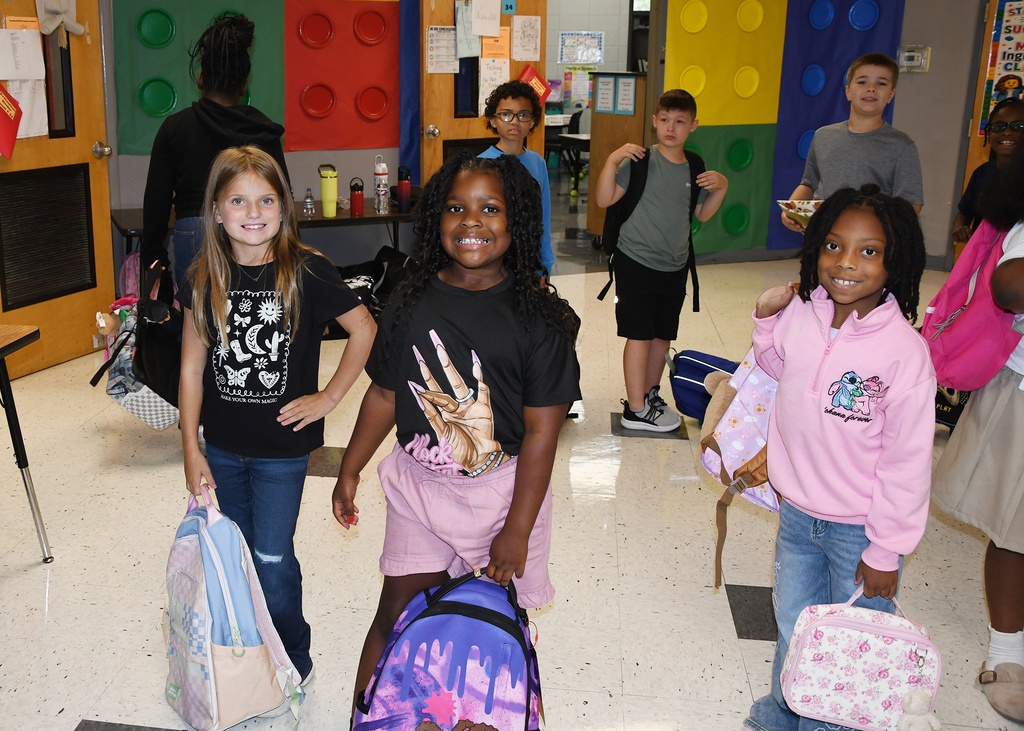 Children in a hallway with colorful block walls, carrying backpacks and holding hands. Some wear jackets.
