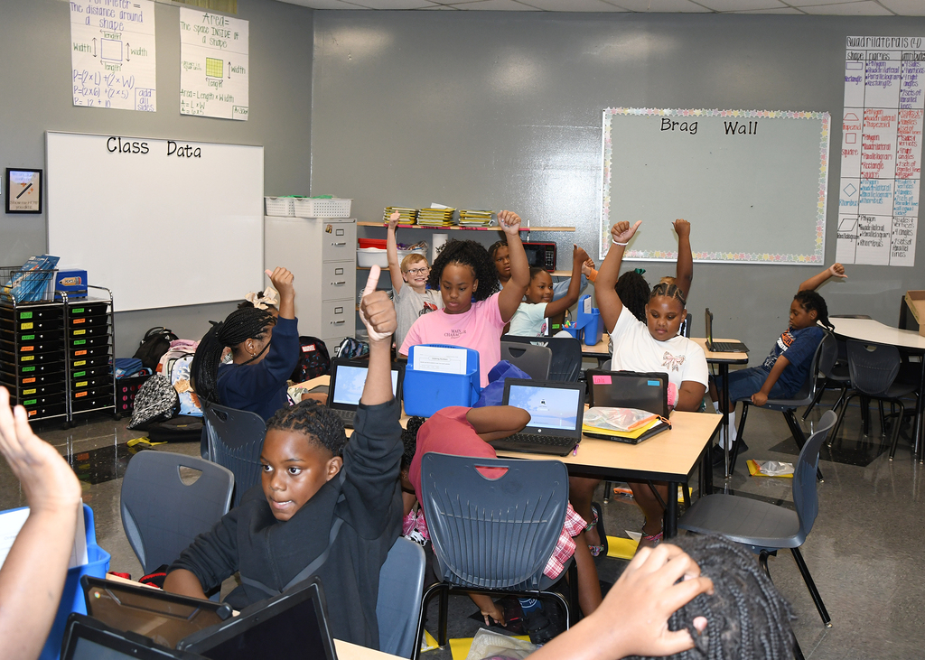A classroom with students seated at desks, raising their hands with laptops. Whiteboards are on the walls.