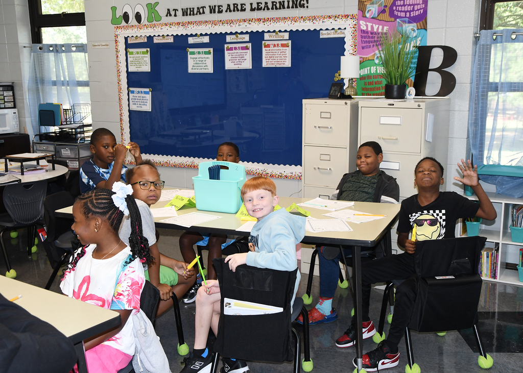Classroom with students seated at tables. A blue bulletin board with announcements is in the background.
