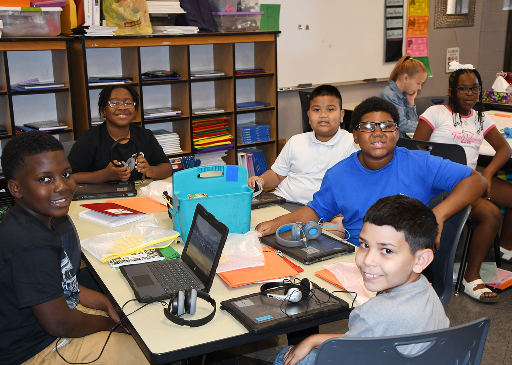 Kids sitting around a table in a classroom with laptops and tablets. Behind them are shelves and a whiteboard.