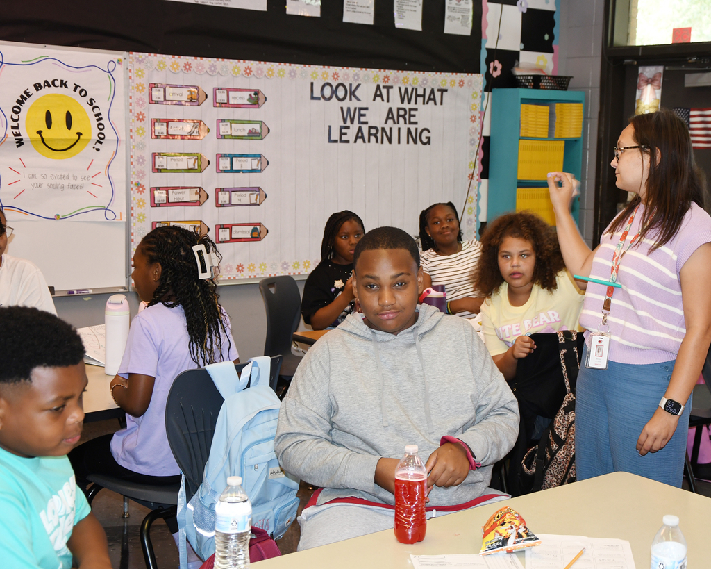 A classroom with a teacher and students. A student sits at a desk, looking at papers.