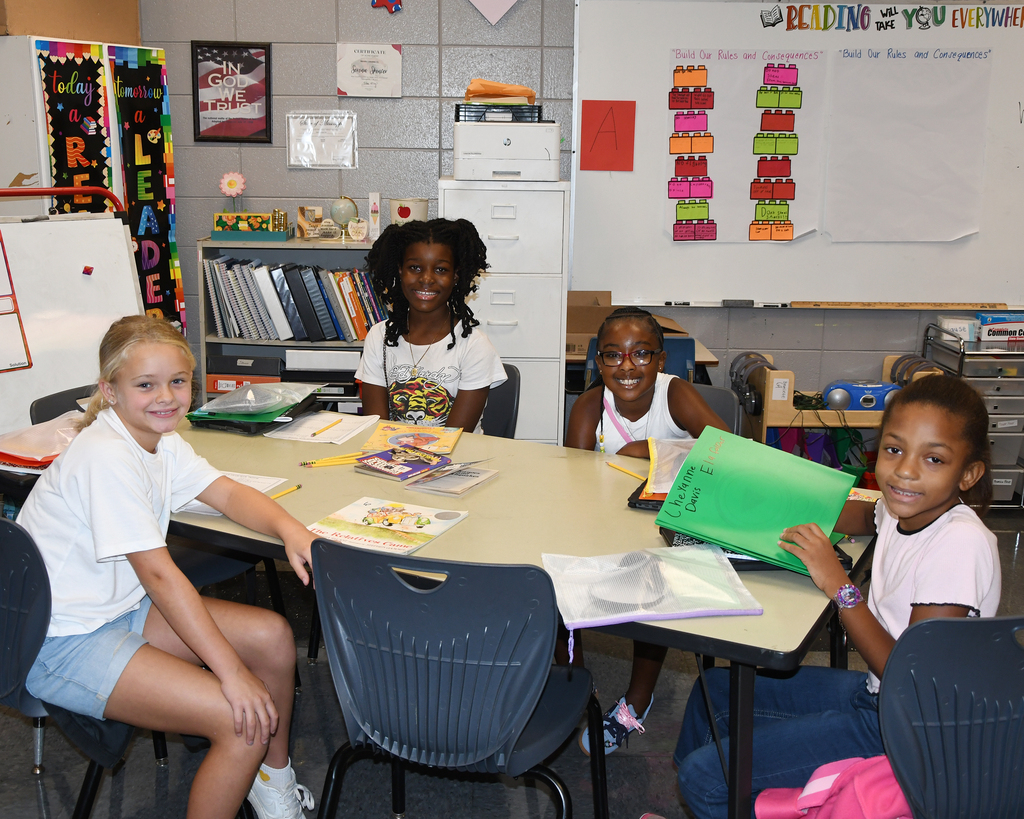 Four children sit at a table in a classroom, smiling. A whiteboard, colorful charts, and a bookshelf are in the background.