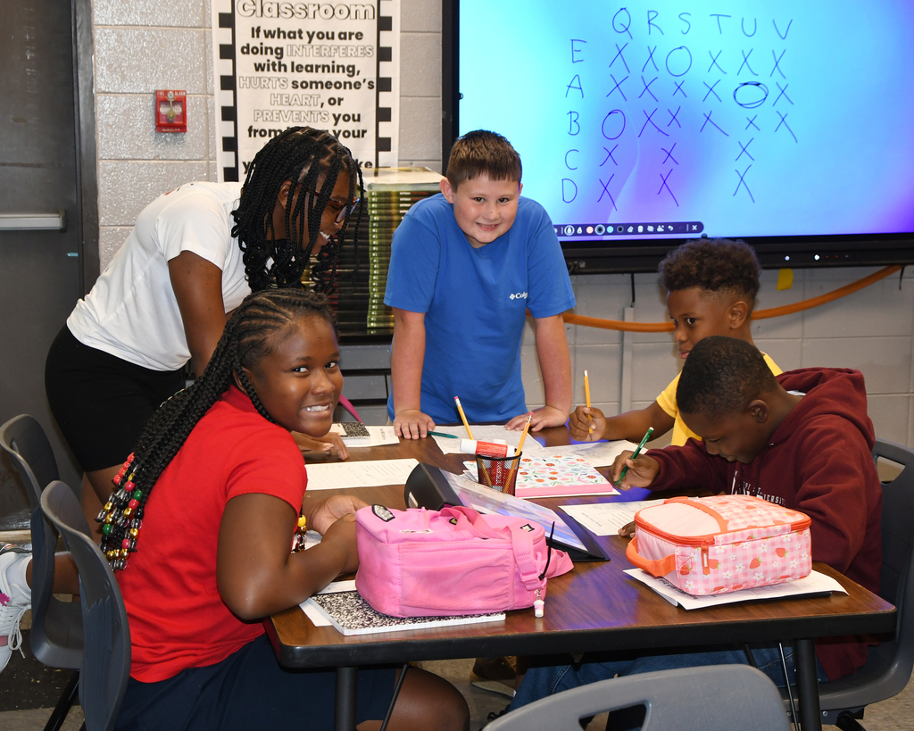 Several children at desks, one with a pink backpack, another with an orange lunchbox, one writing with a pencil.