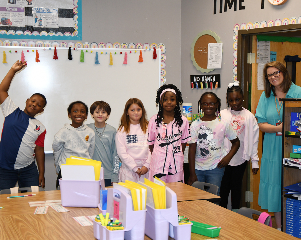A classroom with students standing in front of a whiteboard. A teacher stands to the side.