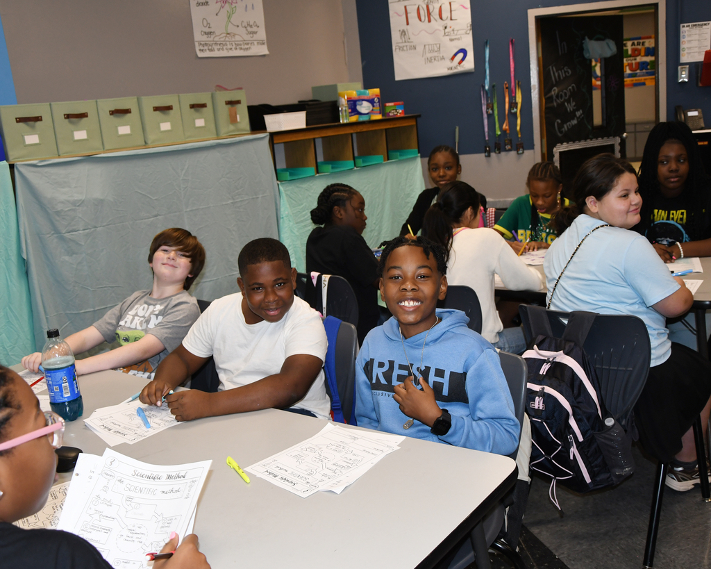 A group of people are seated at tables in a classroom. They have papers and pens in front of them.