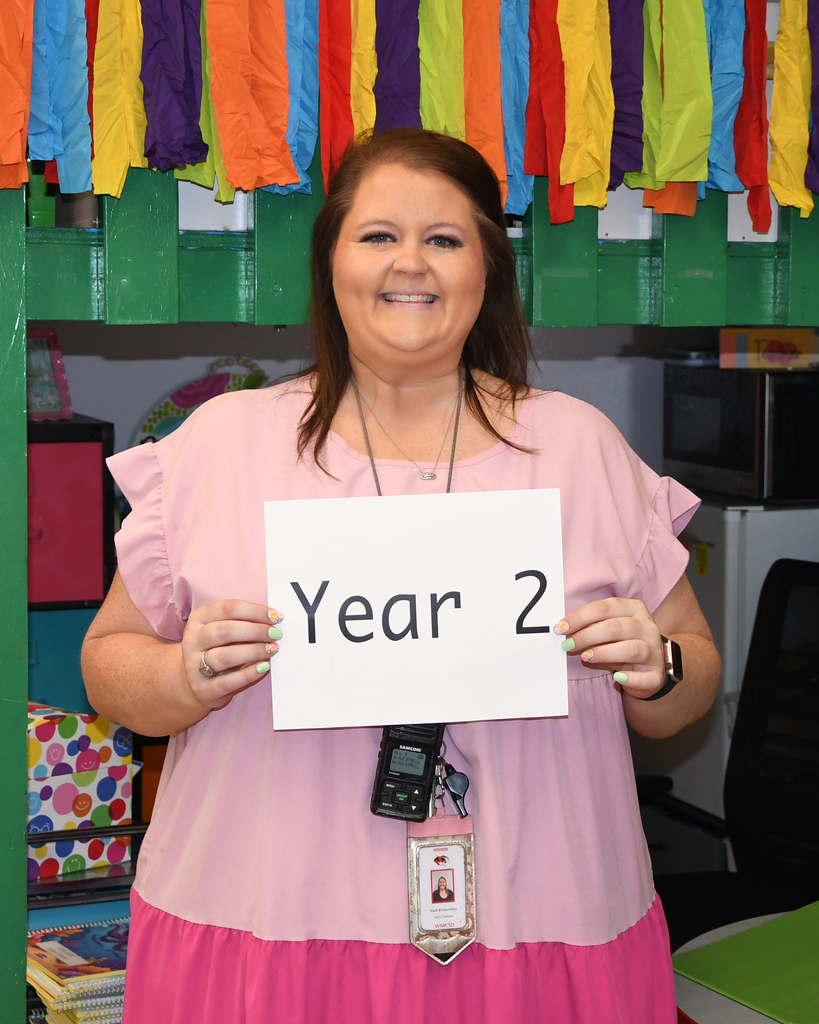 A woman in a pink shirt holds a sign that reads "Year 2" in a classroom.