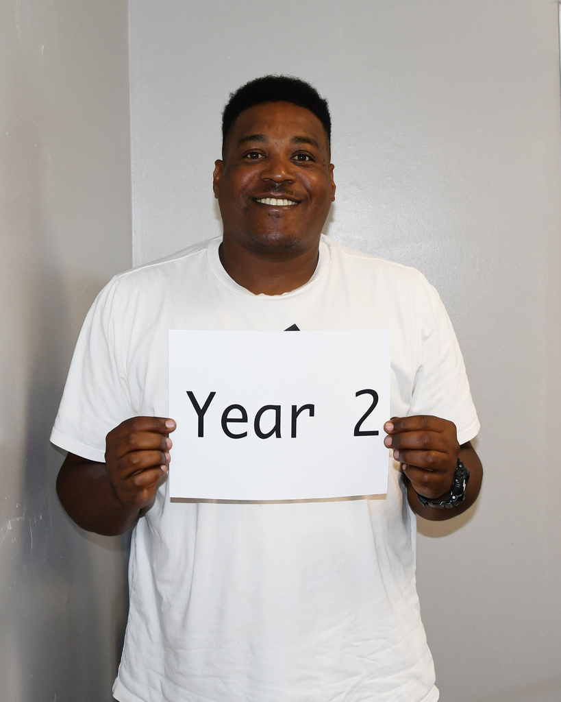 A man in a white shirt holding a sign that reads "Year 2" and smiling, standing against a light gray background.