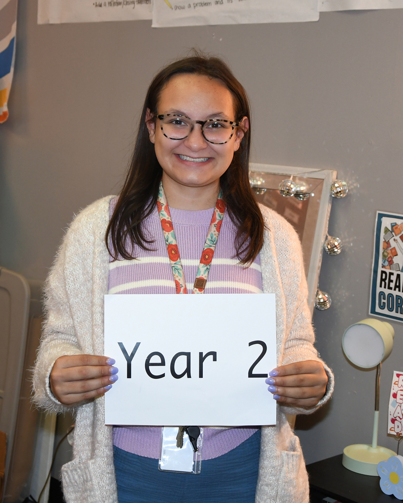 Person holding a sign that says "Year 2" against a gray wall with posters, a lamp, and a framed picture.