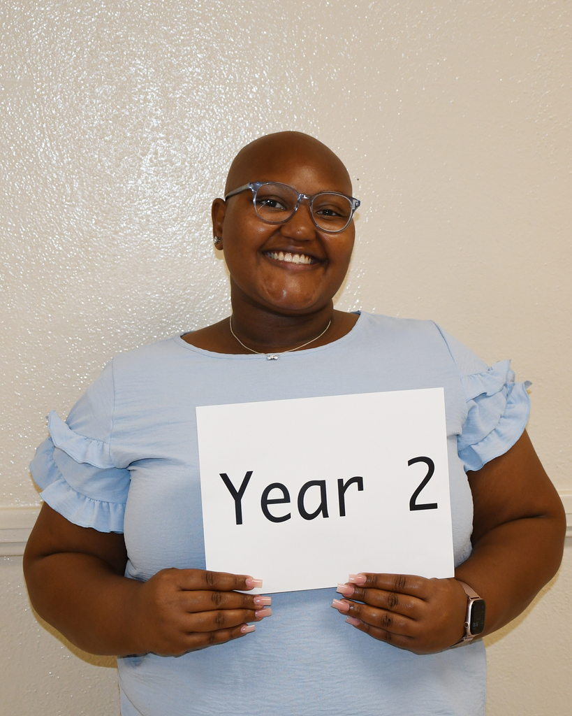 Woman in blue blouse and eyeglasses holding a white sign with black text that reads "Year 2".