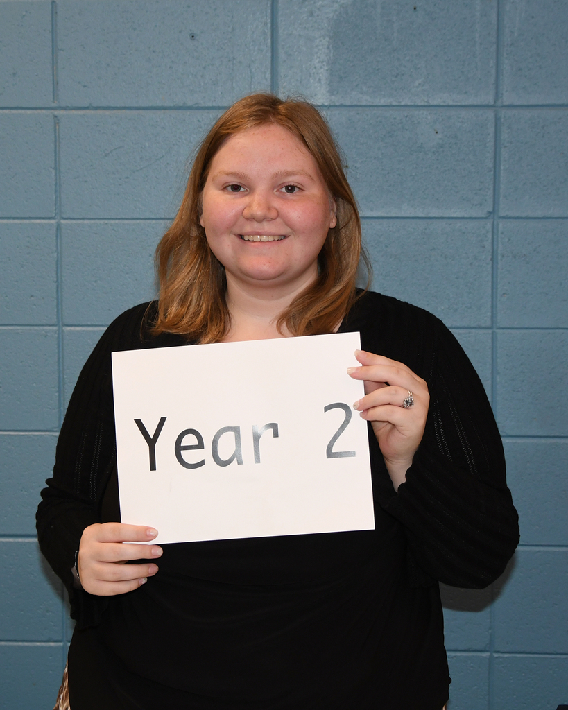 A person smiles while holding a sign that says "Year 2" in front of a blue wall.