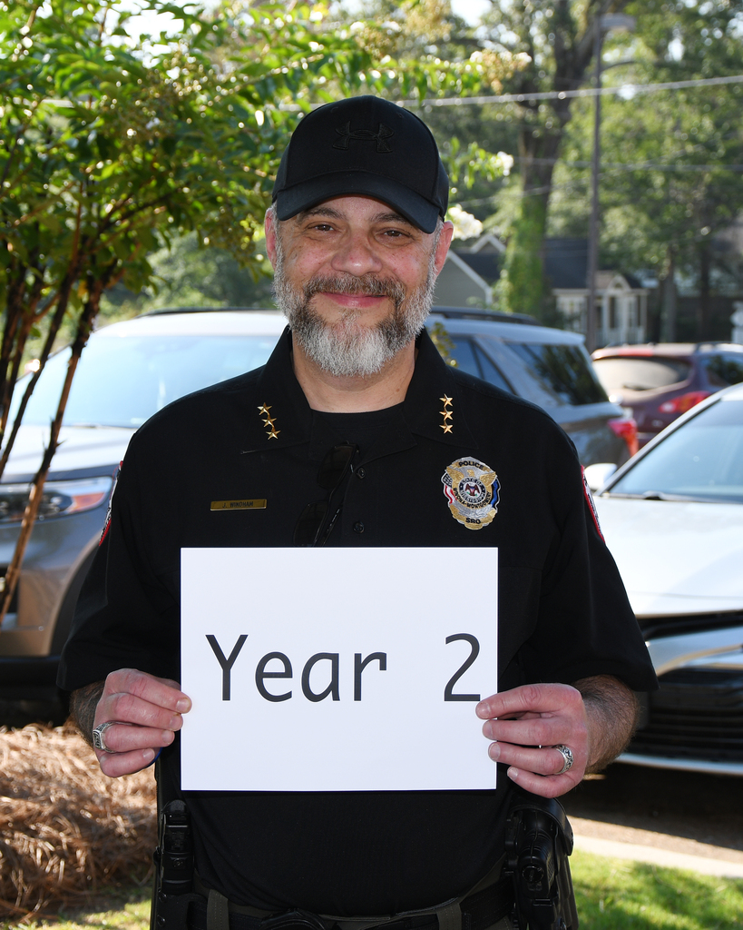 Police officer with a black cap and a uniform holds a sign that says "Year 2." He is smiling.