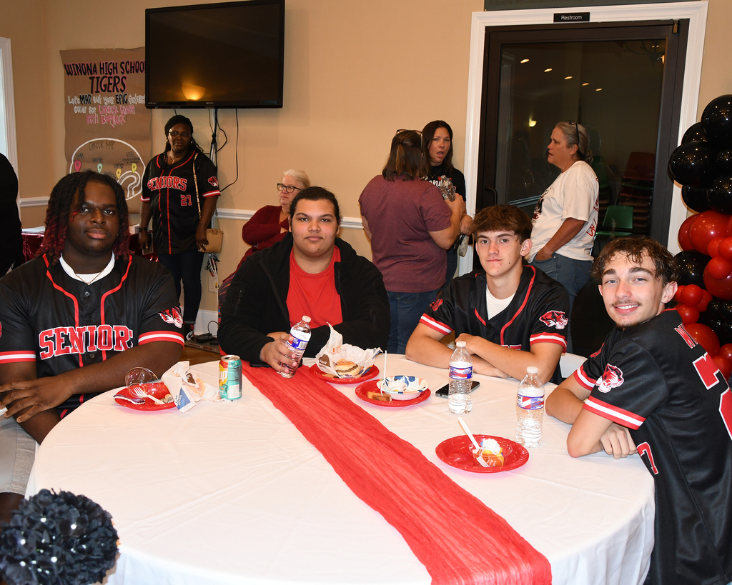 A group of four men at a table with plates, bottles, and a red ribbon. Behind them, people stand in a room with balloons and a television on the wall.