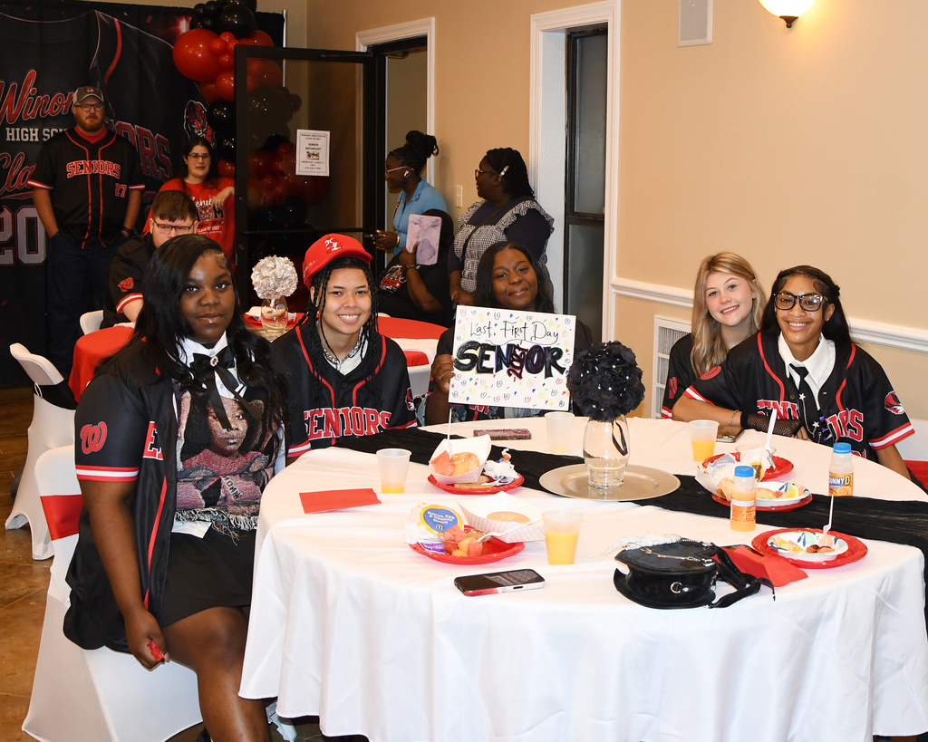A group of six people in red baseball uniforms sit at a table with plates of food.