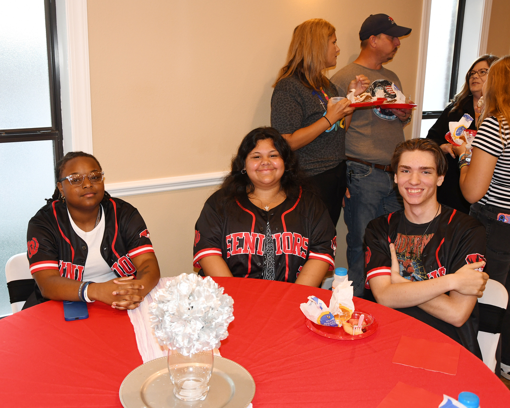 Four people sit at a table wearing baseball jerseys with "SENIORS" printed on the front. One holds a flower centerpiece.