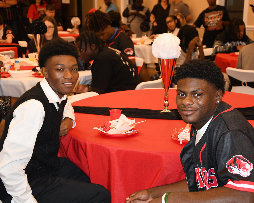 Two young men at a table with a red tablecloth. One wears a white vest, the other a black jersey.