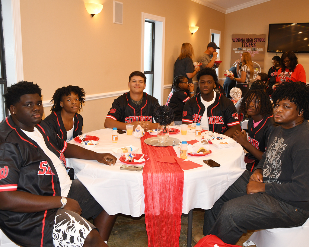 A group of people in red and black jerseys sit at a table with food and drinks in a room.